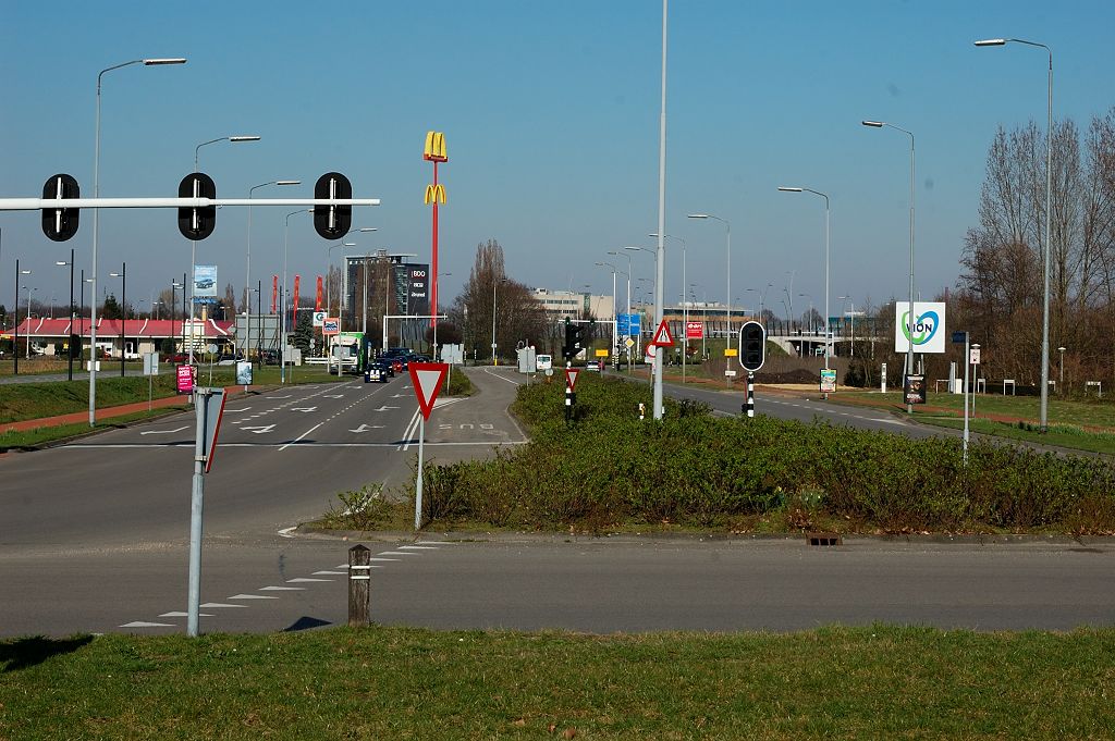 20110320-153729.JPG - Drukste relaties gaan over de Noord-Brabantlaan: naar de N2 (viaduct in de aansluiting nog net zichtbaar) en naar Eindhoven. Het is duidelijk dat hier de verkeersdruk flink kan verminderen als de aansluiting op de N2 ontkoppeld is. In het midden een in onbruik geraakte busbaan, die is verlegd naar het noorden (links). Die ruimte komt mooi van pas voor opstelstroken als de rotonde is omgebouwd tot kruising. Overigens kunnen de verkeerslichten bij de rotonde de weggebruiker op het verkeerde been zetten, om een tweetal redenen. Ten eerste is de beveiliging niet compleet: enkel de loodrecht kruisende hoofdstromen zijn geregeld. Wie de rotonde driekwart neemt moet er dus op bedacht zijn dat hij voorrang moet verlenen aan oprijdend verkeer.